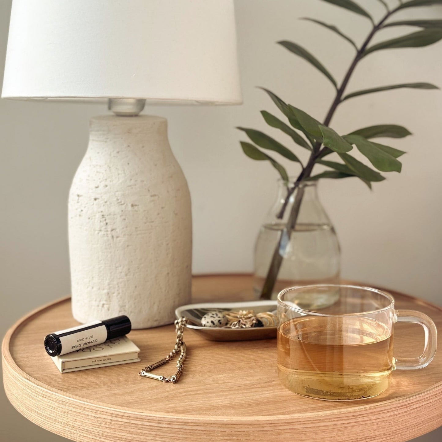 Wooden bedside table with lamp. Plant in vase, jewellery tray, perfume and Naturally Healthful wellness tea on top.