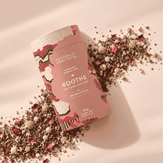 A cylindrical tin of Naturally Healthful Soothe Organic Herbal Tea is styled diagonally on a warm beige background, surrounded by scattered loose herbal tea and dried flower petals.