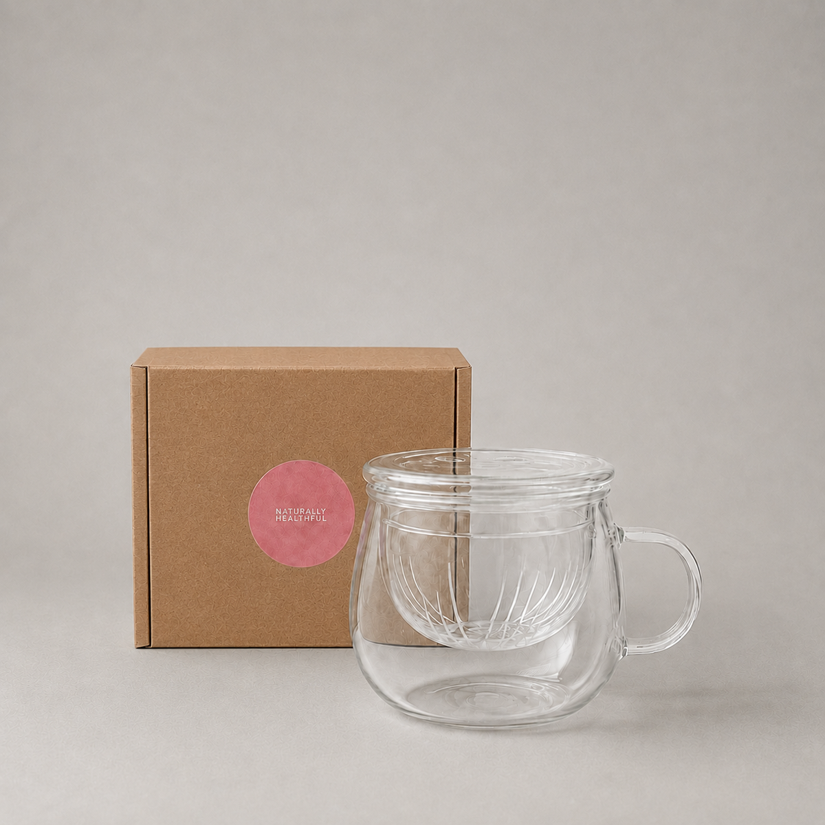 A clear double-walled glass mug with a handle and integrated lid, shown in front of a brown kraft paper box with a pink circular label on a light gray background.