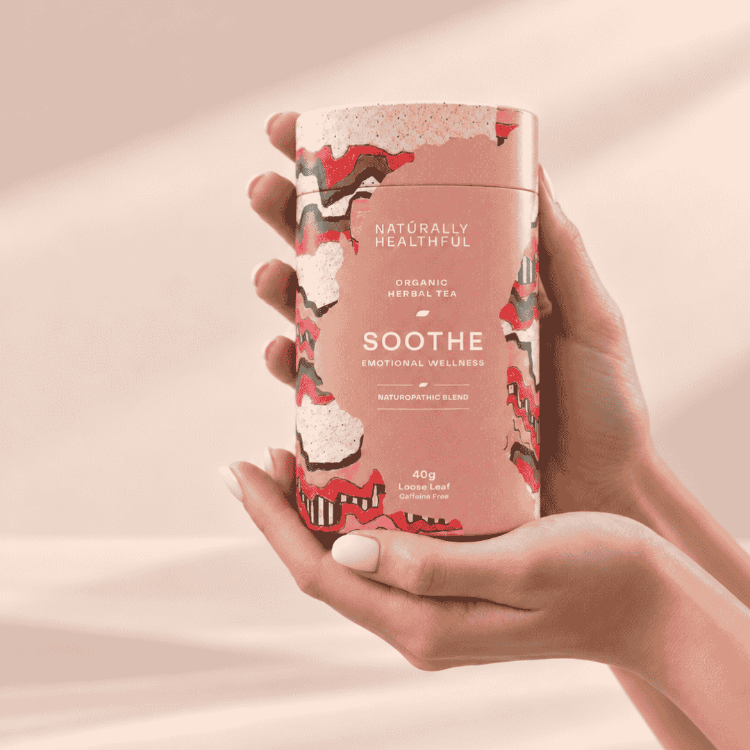 A person's hands holding a cylindrical tin of Naturally Healthful Soothe Emotional Wellness herbal tea against a soft, warm beige background.