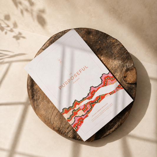 A printed card or booklet titled 'PURPOSEFUL' with a white background and abstract coral, orange, and green line art, placed on a round rustic wooden slice. The scene is styled on a warm beige surface with soft window shadow overlays.