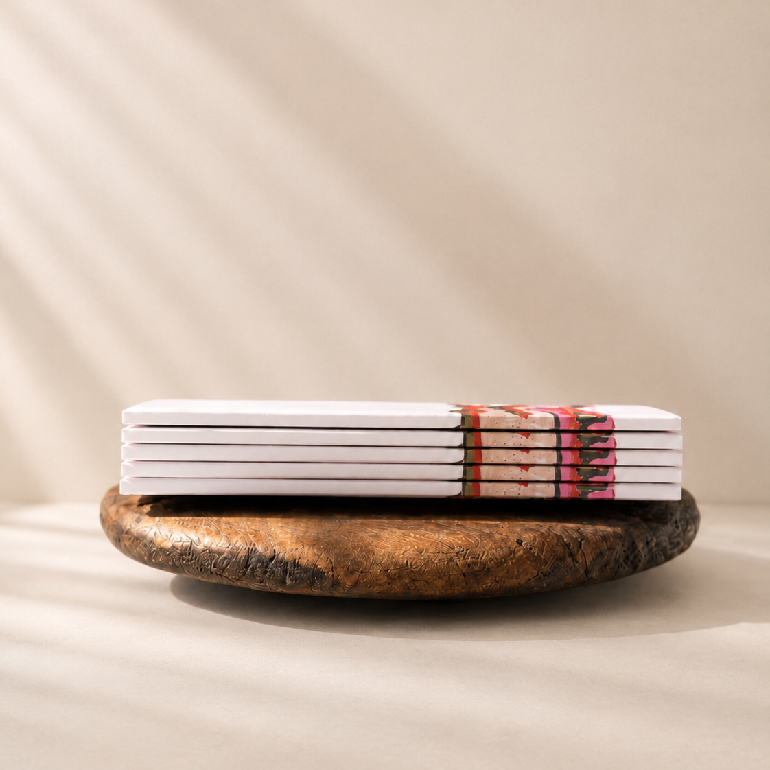 A stack of white rectangular cards or booklets tied with a patterned band, resting on a round rustic wooden board against a warm beige background with soft diagonal shadows.