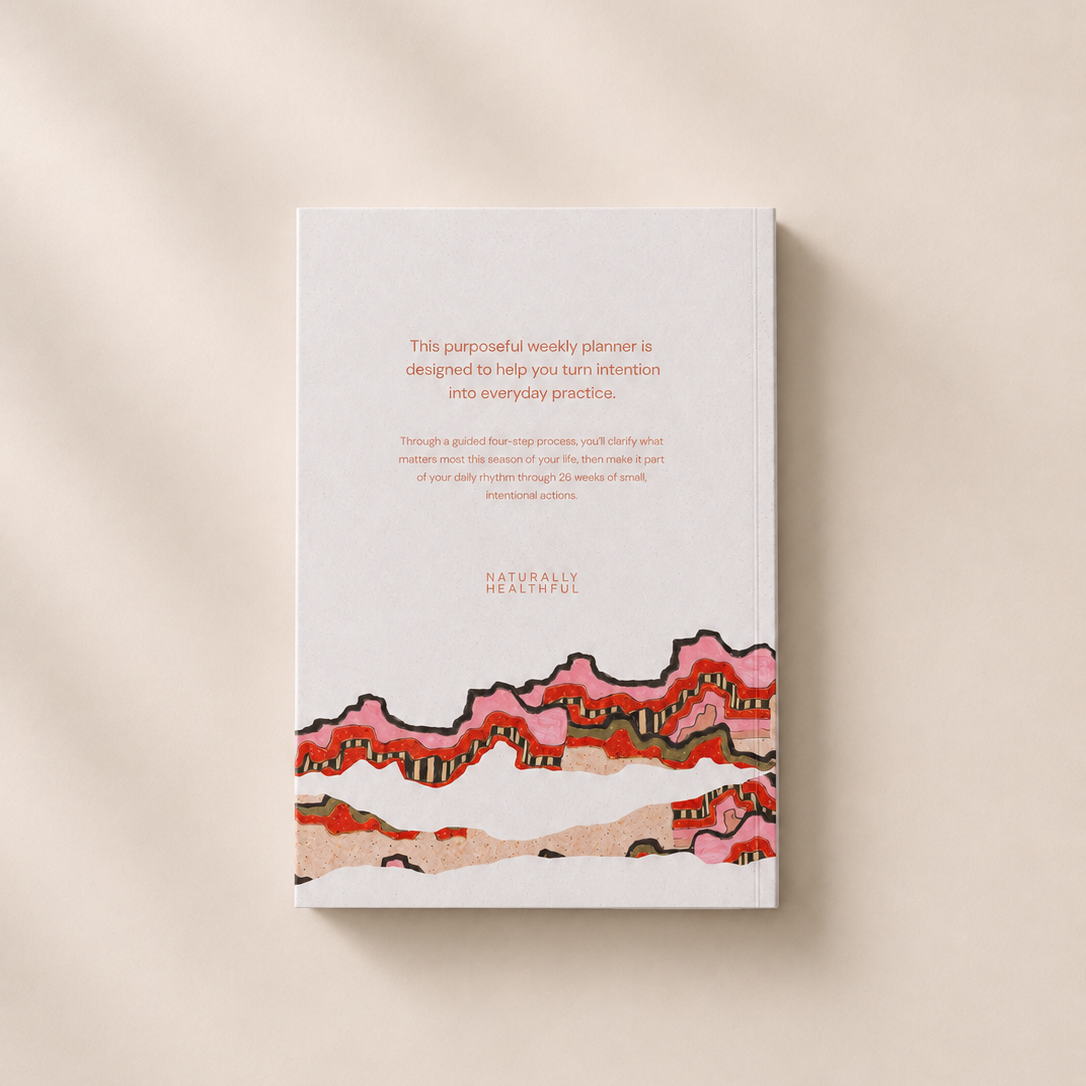 A softcover journal titled 'Natural Balance' lies flat on a warm beige surface with soft, diagonal shadows. The cover is white with a colorful illustration of pink, red, and orange wavy hills at the bottom and small brown text in the center.