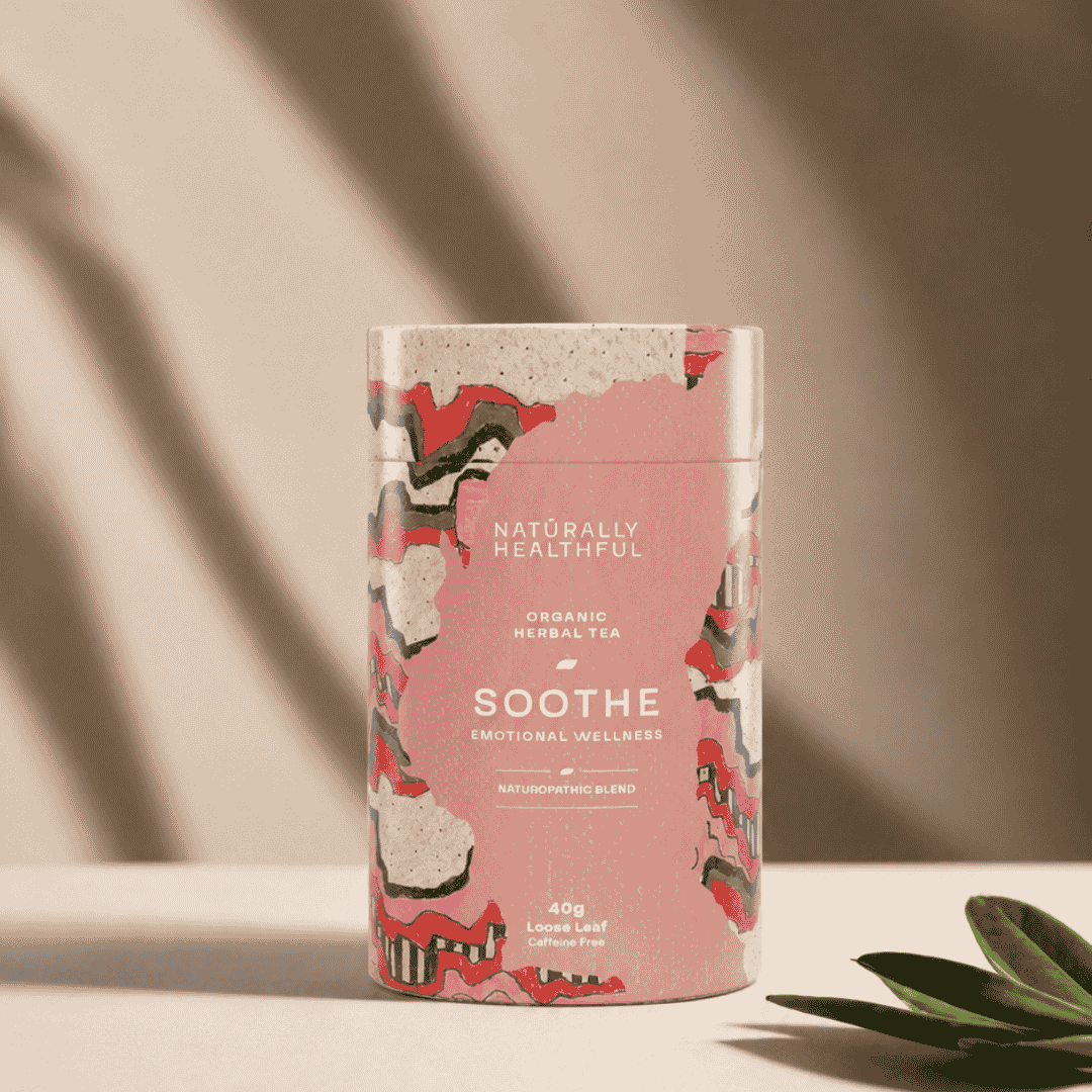 A cylindrical tin of Naturally Healthful Soothe Organic Herbal Tea, featuring a pink label with abstract red, black, and gray patterns, set on a light beige surface with soft shadows and a green leaf in the foreground.